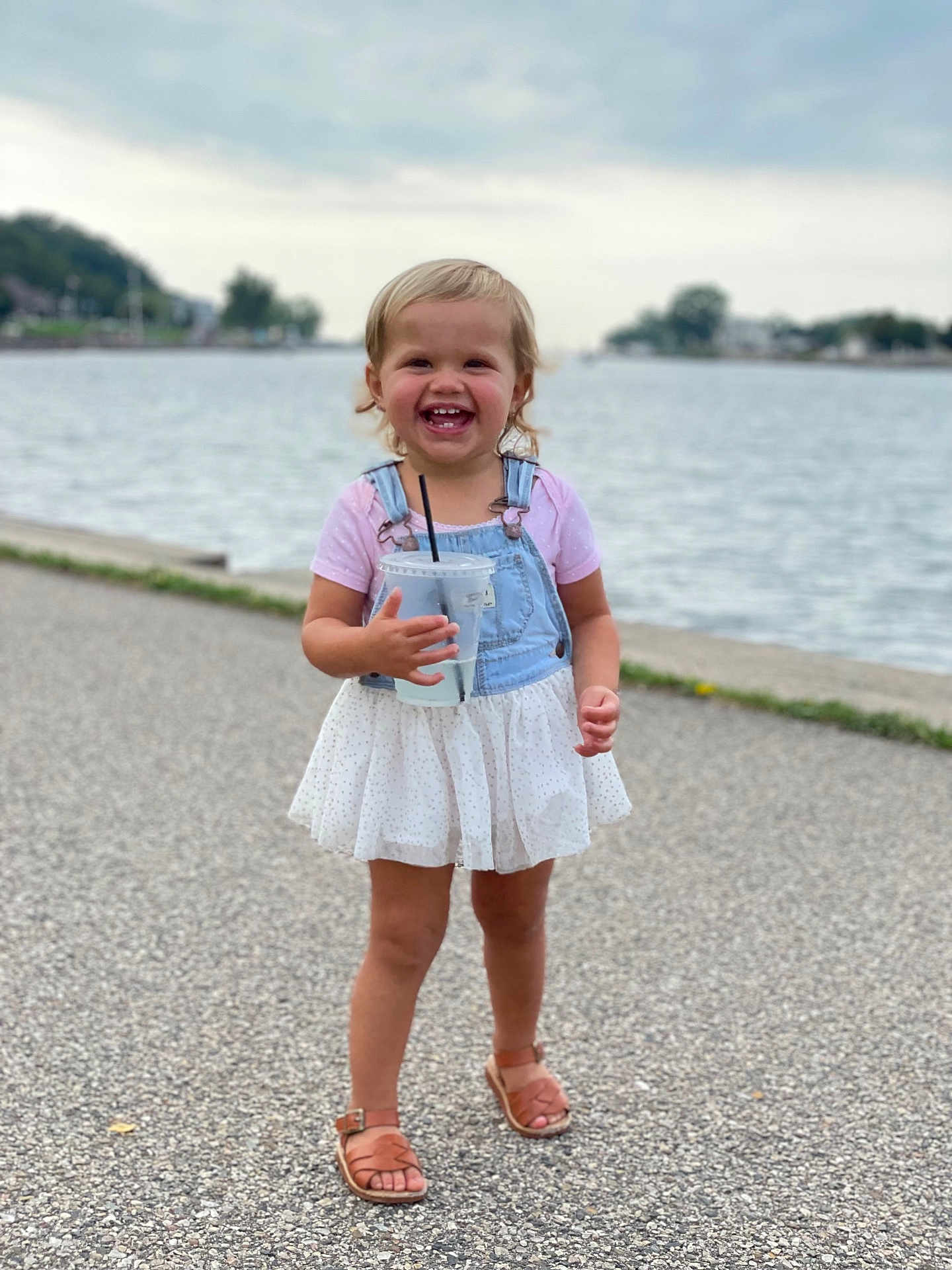 Elly May is registered to the contest to win money with this photo: toddler, child, smiling, outdoor, water, body_of_water, path, sandals, dress, denim, overalls, cup, straw, happy, blonde_hair, short_hair, summer, daytime, nature, portrait