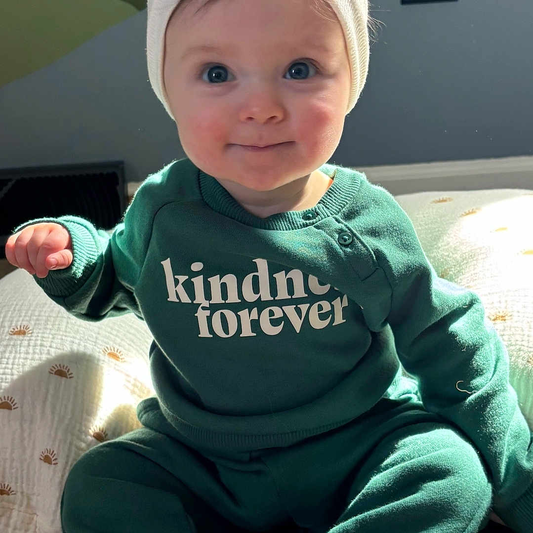 Bailey Rain is registered to the contest to win money with this photo: apparel, baby, barefoot, bow, child, cozy, cute, face, green_clothing, happy, headband, indoor, infant, portrait, quilt, rug, sitting, smiling, sunlight, young_child