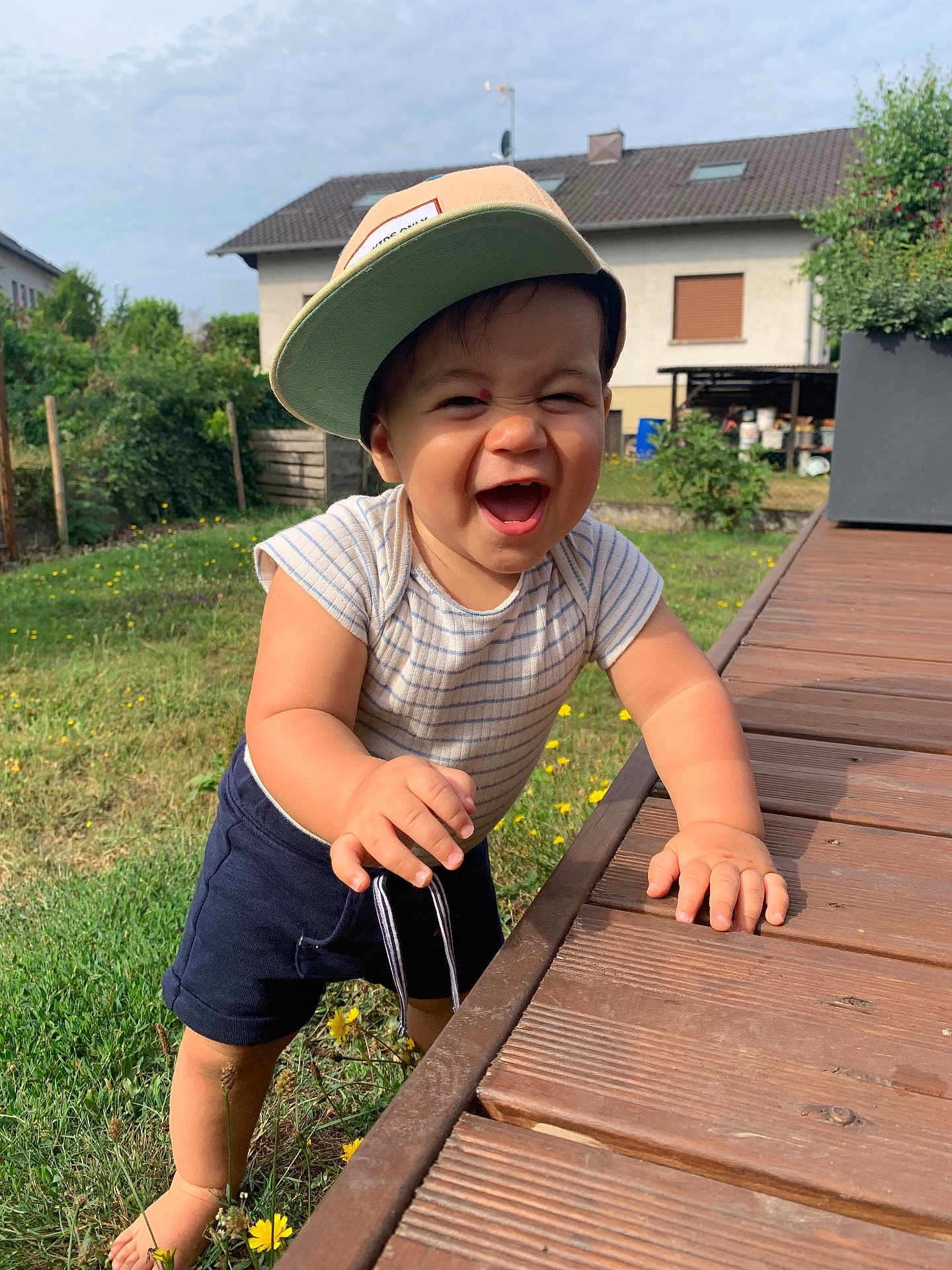 Marius participe au concours pour gagner de l'argent avec cette photo : toddler, child, smiling, cap, striped_shirt, shorts, barefoot, wooden_deck, grass, flowers, garden, house, outdoor, daylight, happy, person, young_child, playful, summer, nature
