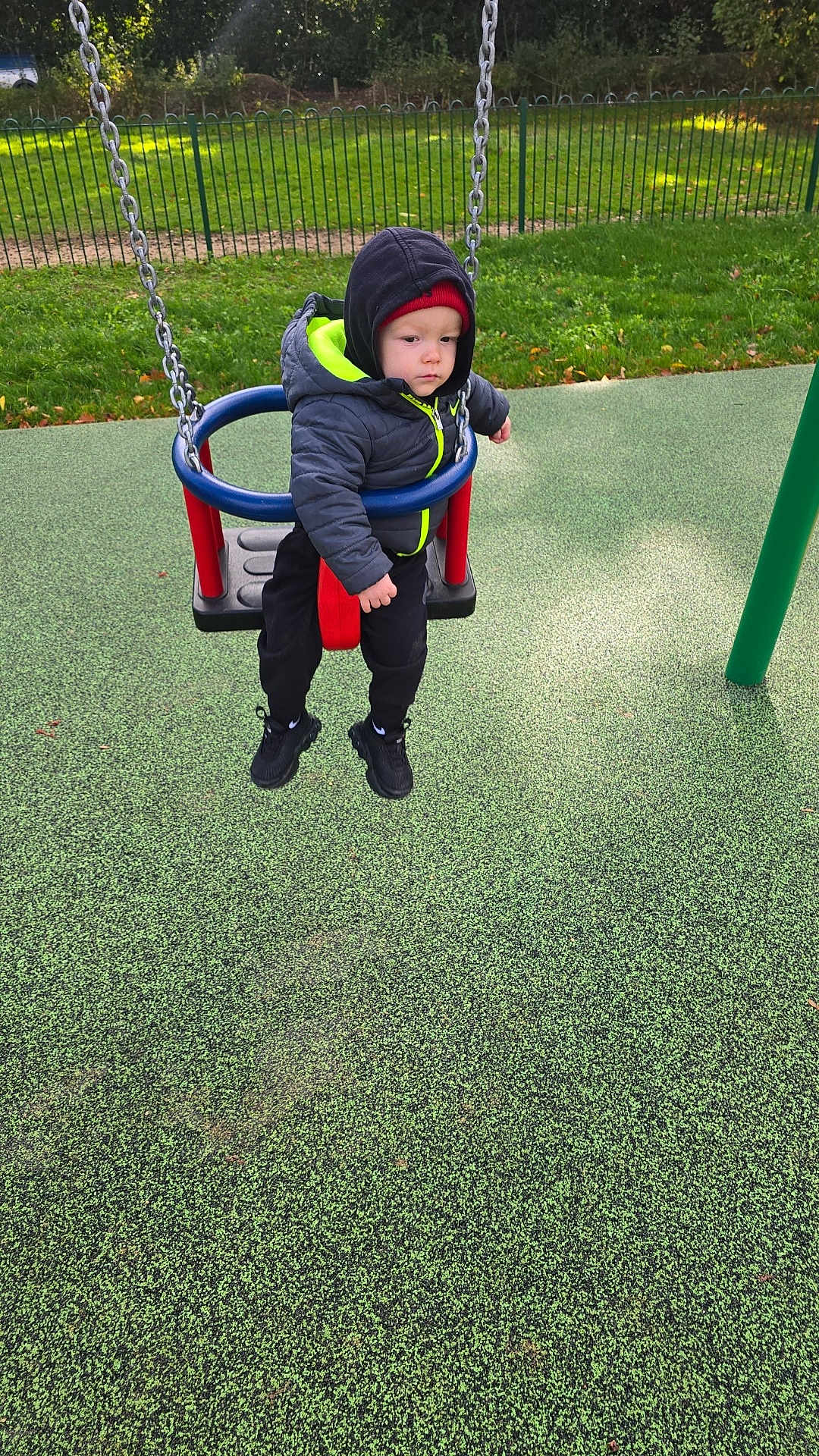 Boston is registered to the contest to win money with this photo: toddler, child, swing, playground, jacket, beanie, outdoor, fence, grass, safety_surface, chain, red, blue, black_shoes, cold_weather, park, person, daytime, serious_expression, playing