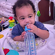 Ares is registered to the contest to win money with this photo: toddler, baby, curly_hair, baby_walker, bottle, toy, indoor, child, person, blue_shirt, plastic, feeding, playtime, cute, young_child, infant, sitting, smiling, home, nursery