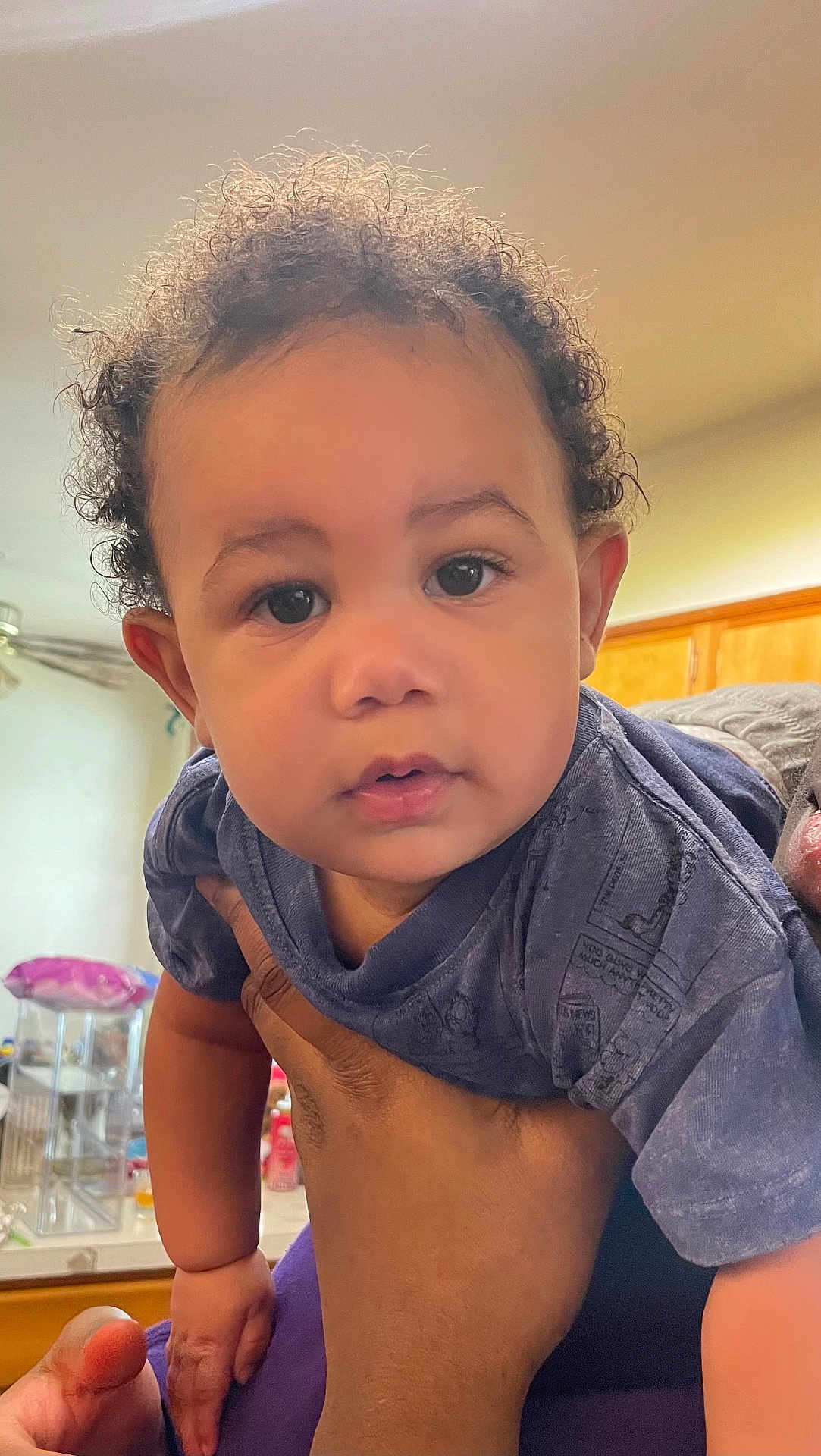 Ares is registered to the contest to win money with this photo: toddler, child, curly_hair, face, hand, indoor, kitchen, person, holding, blue_shirt, expression, cute, baby, skin, eyes, fingertips, furniture, cabinet, light, portrait