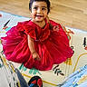 child, toddler, red_dress, smiling, sitting, play_mat, floor, wood_floor, indoor, person, happy, cute, baby, clothing, footwear, home, colorful, portrait, bright, young