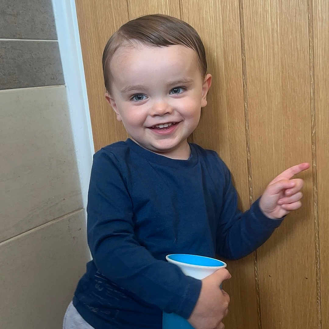 Elijah-Grey joined the competition — help win amazing prizes! blue_cup, casual_clothing, child, cute, expression, face, gray_pants, hair, happy, indoor, light, long_sleeve_shirt, person, playful, pointing, skin, smiling, standing, toddler, wooden_door