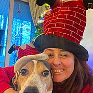 Blue Angel is registered to the contest to win money with this photo: woman, dog, christmas_hat, holiday, indoor, smile, pet, festive, tree, lights, decorations, red_clothing, face, closeup, hug, cheerful, window, curtains, seasonal, funny_hat