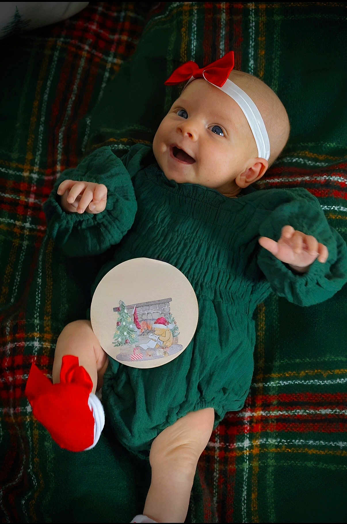 Layla joined the competition — help win amazing prizes! baby, infant, smiling, green_dress, red_bow, red_socks, headband, plaid_blanket, holiday, christmas_theme, cute, happy, child, portrait, indoors, cozy, laying_down, festive, person, celebration