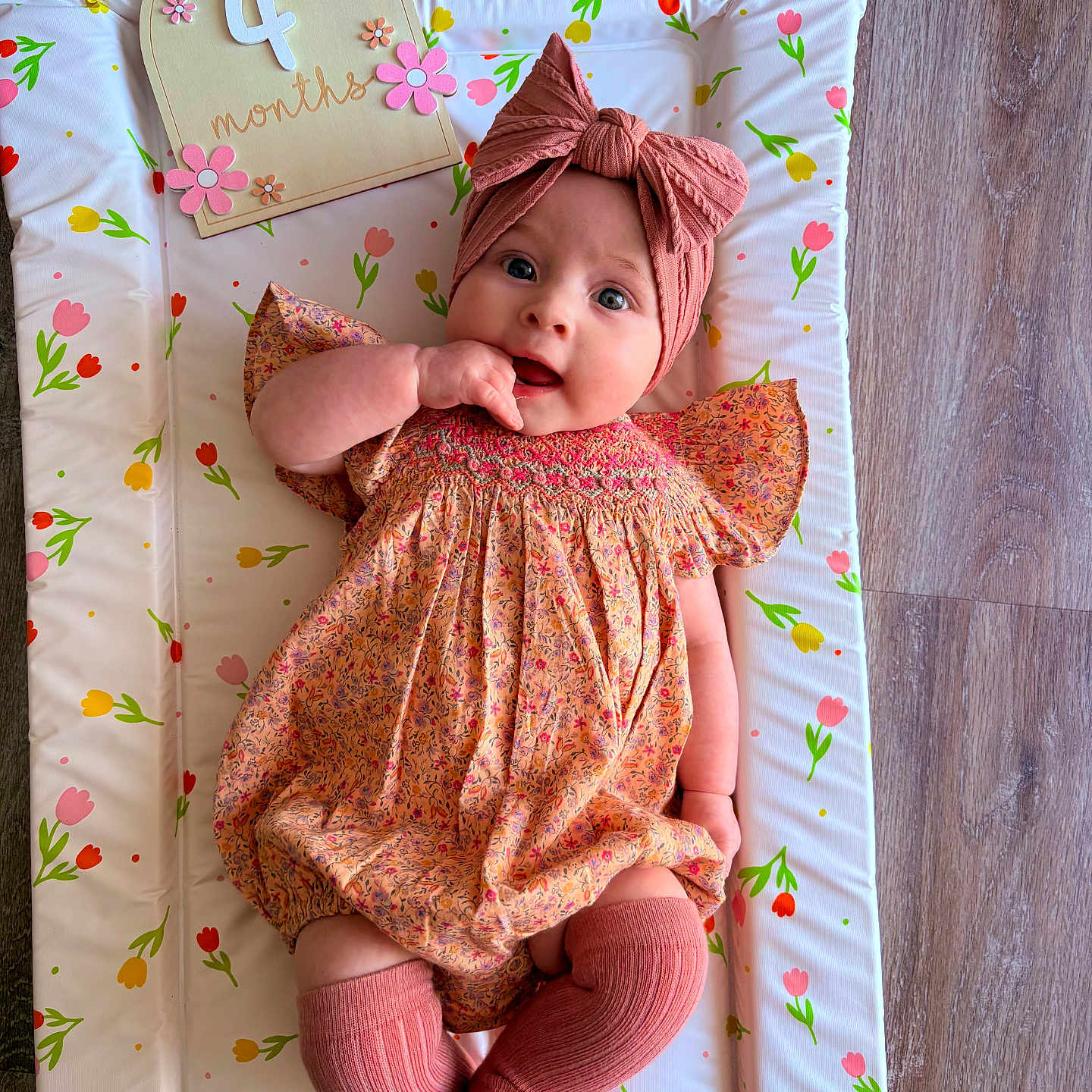 Mabel-Blossom is registered to the contest to win money with this photo: 4_months, baby, changing_pad, child, cute, dress, flooring, floral_pattern, hand_on_mouth, happy, headband, indoors, infant, knee_socks, milestone, orange, person, pink, portrait, wooden_sign
