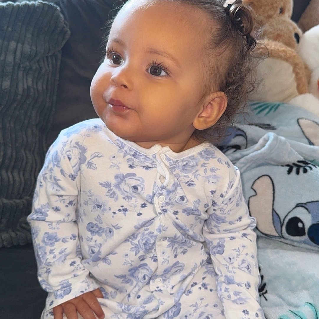 River-Lea is registered to the contest to win money with this photo: baby, blanket, child, couch, cozy, curly_hair, cute, face, floral_pattern, furniture, hand, home, indoor, onesie, plush_toy, portrait, sitting, smiling, soft_lighting, toddler