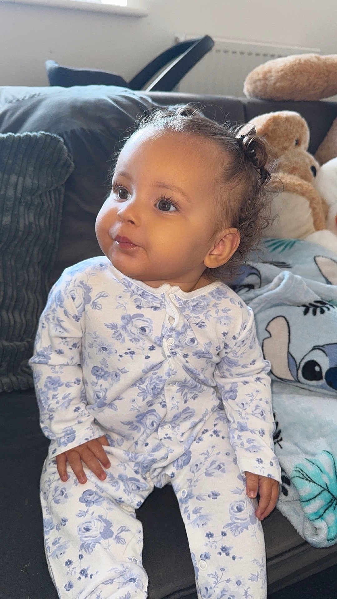 River-Lea is registered to the contest to win money with this photo: toddler, child, baby, onesie, floral_pattern, couch, plush_toy, blanket, indoor, curly_hair, sitting, portrait, cute, soft_lighting, cozy, smiling, face, hand, furniture, home