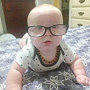 Cotton is registered to the contest to win money with this photo: baby, glasses, bed, blanket, pattern, star_pattern, necklace, infant, clothing, face, hand, room, furniture, dresser, curious, lying_down, indoors, portrait, child, cute