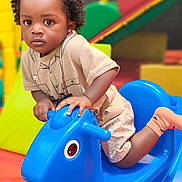 Shad a rejoint le concours — aidez-le/la à gagner de superbes lots ! toddler, child, toy, rocking_horse, blue, indoor, play_area, colorful, plastic, socks, shorts, shirt, curly_hair, face, hands, eyes, floor, background, playground_equipment, young_child