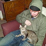 Trouble is registered to the contest to win money with this photo: man, cat, indoor, chair, hoodie, beanie, smartphone, wooden_cabinet, candles, radio, striped_chair, tabby_cat, carpet, smiling, casual_clothing, relaxed, furniture, home_decor, person, pet