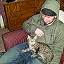 man, cat, indoor, chair, hoodie, beanie, smartphone, wooden_cabinet, candles, radio, striped_chair, tabby_cat, carpet, smiling, casual_clothing, relaxed, furniture, home_decor, person, pet