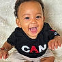 baby, smiling, child, black_tshirt, beige_shorts, curly_hair, sitting, fluffy_blanket, happy, portrait, indoor, cute, joyful, young_child, hand_bracelet, bright_eyes, open_mouth, skin, person, closeup