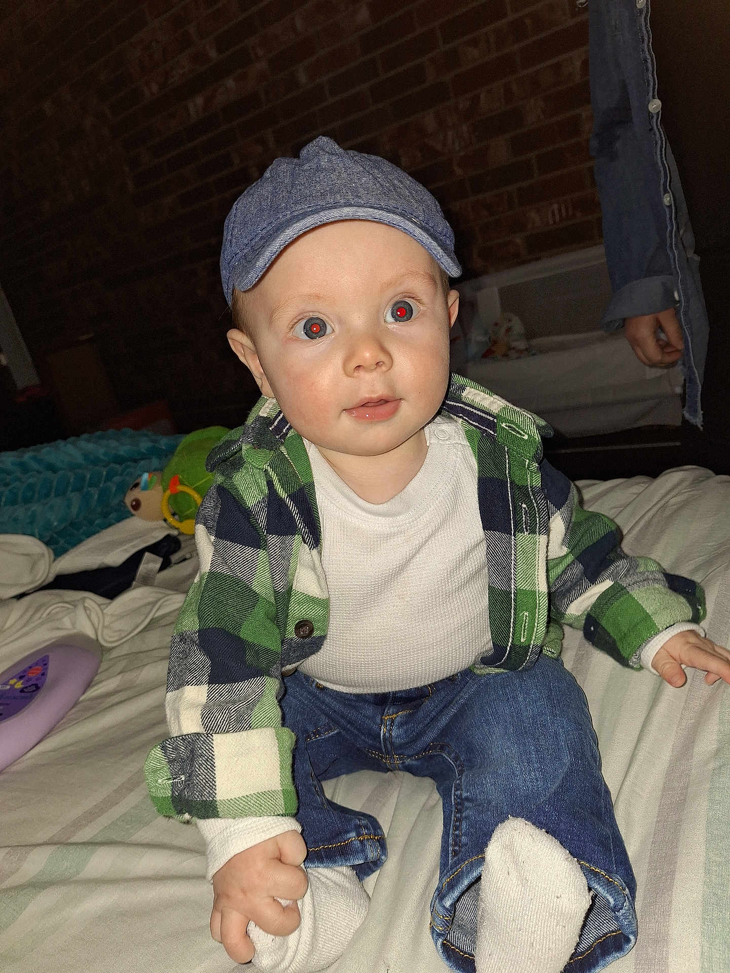 Nazareth is registered to the contest to win money with this photo: baby, child, cap, plaid_shirt, white_shirt, denim_jeans, socks, bed, brick_wall, indoor, person, cute, clothing, curious_expression, hand, toy, blanket, casual, portrait, infant