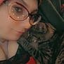 affection, casual_clothing, cat, close_up, cozy, cute, eyewear, face, glasses, hat, indoor, knit_hat, person, portrait, resting, sleepy_cat, snuggling, tabby_cat, tattoo, warm