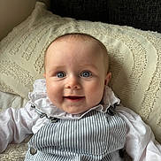 Matilda is registered to the contest to win money with this photo: baby, child, smiling, blue_eyes, face, portrait, white_shirt, striped_overall, pillow, blanket, cozy, indoors, cute, happy, infant, young_child, soft_lighting, texture, clothing, person