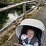 Matilda is registered to the contest to win money with this photo: baby, stroller, blanket, outdoor, bridge, water, nature, greenery, child, pathway, wooden_fence, smiling, happy, person, infant, blue_eyes, clothing, canopy, hand, park
