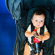 Yassine a rejoint le concours — aidez-le/la à gagner de superbes lots ! baby, black_stroller, blue_background, child, clothing, cute, expression, floor_tiles, hand, holding, indoors, infant, person, red_cap, seat, sitting, soda_bottle, straps, stroller, young_child