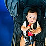 baby, black_stroller, blue_background, child, clothing, cute, expression, floor_tiles, hand, holding, indoors, infant, person, red_cap, seat, sitting, soda_bottle, straps, stroller, young_child