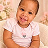 baby, background_blur, child, cozy, curly_hair, cute, cute_expression, face, flowers, hands, happy, indoor, pink_clothing, portrait, sitting, smiling, soft_furniture, toddler, warm_lighting, young_child