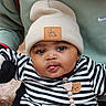 baby, child, face, beanie, hat, striped_clothing, black_and_white, sweater, tongue_out, cute, adorable, person, infant, earring, outdoor, holding, green_sweatshirt, brown_strap, playful, portrait