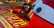 Denim is registered to the contest to win money with this photo: toddler, child, car, toy_car, red, black_clothing, durag, rust_eze, gas_pumps, outdoor, sunlight, vintage, smiling, sitting, cartoon_eyes, holding, vehicle, playful, portrait, daylight