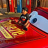 toddler, child, car, toy_car, red, black_clothing, durag, rust_eze, gas_pumps, outdoor, sunlight, vintage, smiling, sitting, cartoon_eyes, holding, vehicle, playful, portrait, daylight