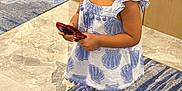 Olive is registered to the contest to win money with this photo: blue, carpet, child, curly_hair, cute, dress, floor, footwear, hat, holding, indoor, looking, marble, patterned_clothing, person, red_object, sandals, standing, toddler, young_child