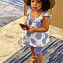 blue, carpet, child, curly_hair, cute, dress, floor, footwear, hat, holding, indoor, looking, marble, patterned_clothing, person, red_object, sandals, standing, toddler, young_child