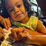 Olive is registered to the contest to win money with this photo: activity_sheet, adult_hand, child, coloring, crayon, curly_hair, dress, education, face, girl, hand, indoor, learning, natural_light, paper, person, plaid, smile, table, window