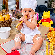 Jumana is registered to the contest to win money with this photo: baby, child, chef_hat, baking, bread, breadsticks, flour, kitchen, indoor, sitting, smiling, eating, food, winnie_the_pooh, stuffed_toy, rolling_pin, basket, table, portrait, playful