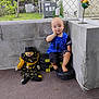 toddler, child, clown_doll, weights, barefoot, blue_shirt, spider_web, concrete_wall, chain_link_fence, carpet, indoor, sitting, curious, toy, corner, yellow, black, plastic, home, portrait