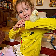 Daisy participe au concours pour gagner de l'argent avec cette photo : child, yellow_shirt, pikachu, chick, pet, living_room, carpet, wooden_furniture, toy_blocks, smiling, curly_hair, portrait, indoor, cozy, family, playful, person, arm, cute, happy