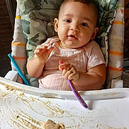 Rayla is registered to the contest to win money with this photo: baby, high_chair, food, spoon, messy, cushion, camouflage, infant, feeding, child, cute, indoor, portrait, face, table, smile, clothing, hands, expression, seat