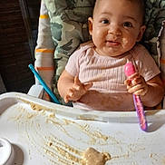 Rayla is registered to the contest to win money with this photo: baby, camouflage, child, clothing, cute, face, feeding, food, hands, happy, high_chair, indoor, infant, meal, messy, pink, portrait, seat, smiling, spoon