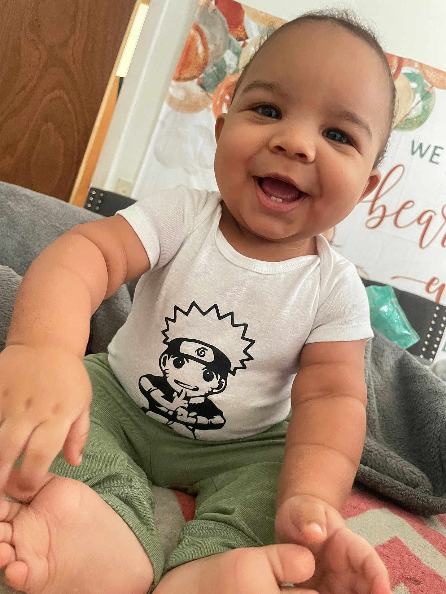 Ezrah joined the competition — help win amazing prizes! baby, child, smile, happy, onesie, cartoon_print, green_pants, sitting, blanket, indoor, face, hand, foot, cozy, cute, infant, portrait, home, decor, playful