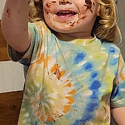 Hunter is registered to the contest to win money with this photo: child, toddler, blonde_hair, curly_hair, chocolate, messy_face, tie_dye_shirt, smiling, happy, cake, food, plate, table, indoor, kitchen, hand, dirt, mess, eating, person