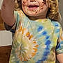 child, toddler, blonde_hair, curly_hair, chocolate, messy_face, tie_dye_shirt, smiling, happy, cake, food, plate, table, indoor, kitchen, hand, dirt, mess, eating, person