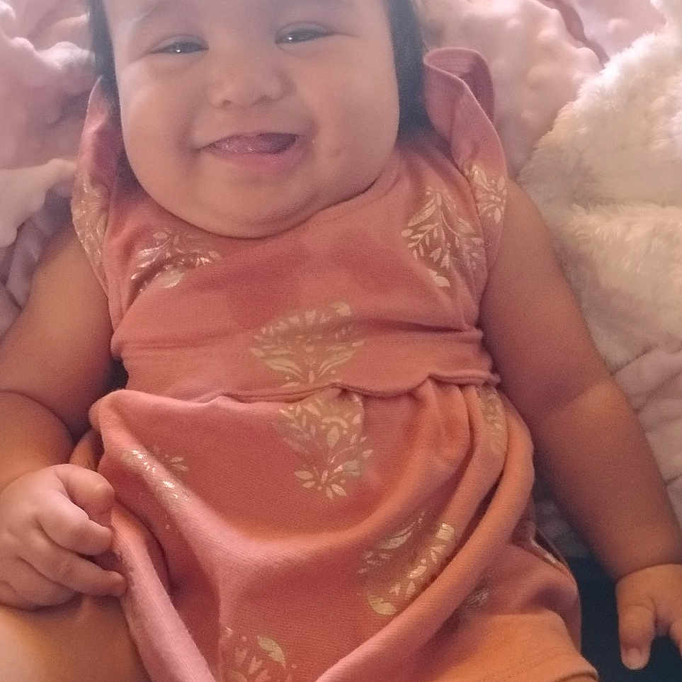 Kauionalani is registered to the contest to win money with this photo: baby, blanket, bow, child, chubby_cheeks, cozy, cute, diaper, face, happy, indoors, infant, person, pink_dress, plush, portrait, sitting, smiling, soft_toys, young_child