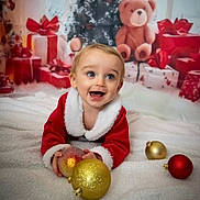 Thomas is registered to the contest to win money with this photo: celebration, child, christmas, christmas_tree, cute, decorations, face, festive, happy, holiday, indoors, ornaments, portrait, presents, red_clothing, smiling, teddy_bear, toddler, toy, white_blanket