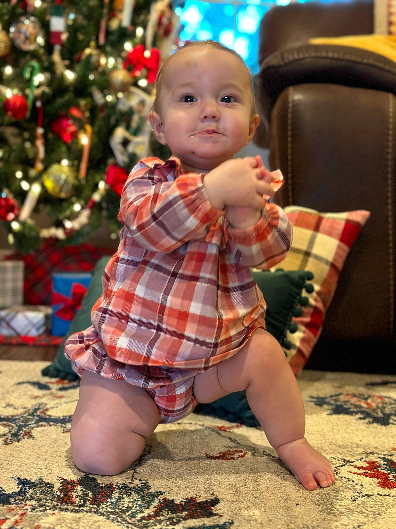 Emilee is registered to the contest to win money with this photo: baby, baby_toddler_clothing, child, christmas_tree, event, eye, face, flooring, fun, happy, human_leg, pattern, people, people_in_nature, person, plaid, sitting, standing, tartan, thigh