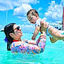 baby, woman, water, ocean, swimming, sunglasses, floral_clothing, sky, clouds, sailboats, sunny, outdoor, playful, happy, lifting, vacation, summer, child, family, fun