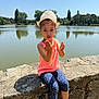 blue_sky, casual_clothing, child, daytime, gesture, girl, hat, lake, leggings, nature, outdoor, pink_shoes, portrait, sitting, stone_wall, summer, sunny, trees, water, young
