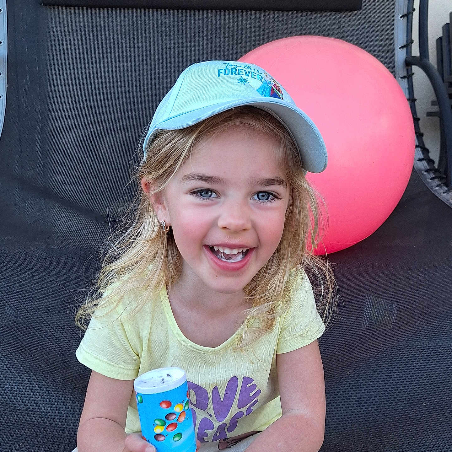 Olivia a rejoint le concours — aidez-le/la à gagner de superbes lots ! child, girl, smiling, blue_eyes, cap, yellow_shirt, white_pants, pink_ball, chair, outdoor, happy, holding_object, playful, blond_hair, casual_clothing, daylight, portrait, sitting, toy, fun