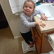 Madison is registered to the contest to win money with this photo: child, smiling, bathroom, sink, step_stool, towel, toiletries, countertop, floor, cabinet, person, barefoot, long_sleeve_shirt, black_pants, hair_tie, happy, indoor, young_child, standing, holding
