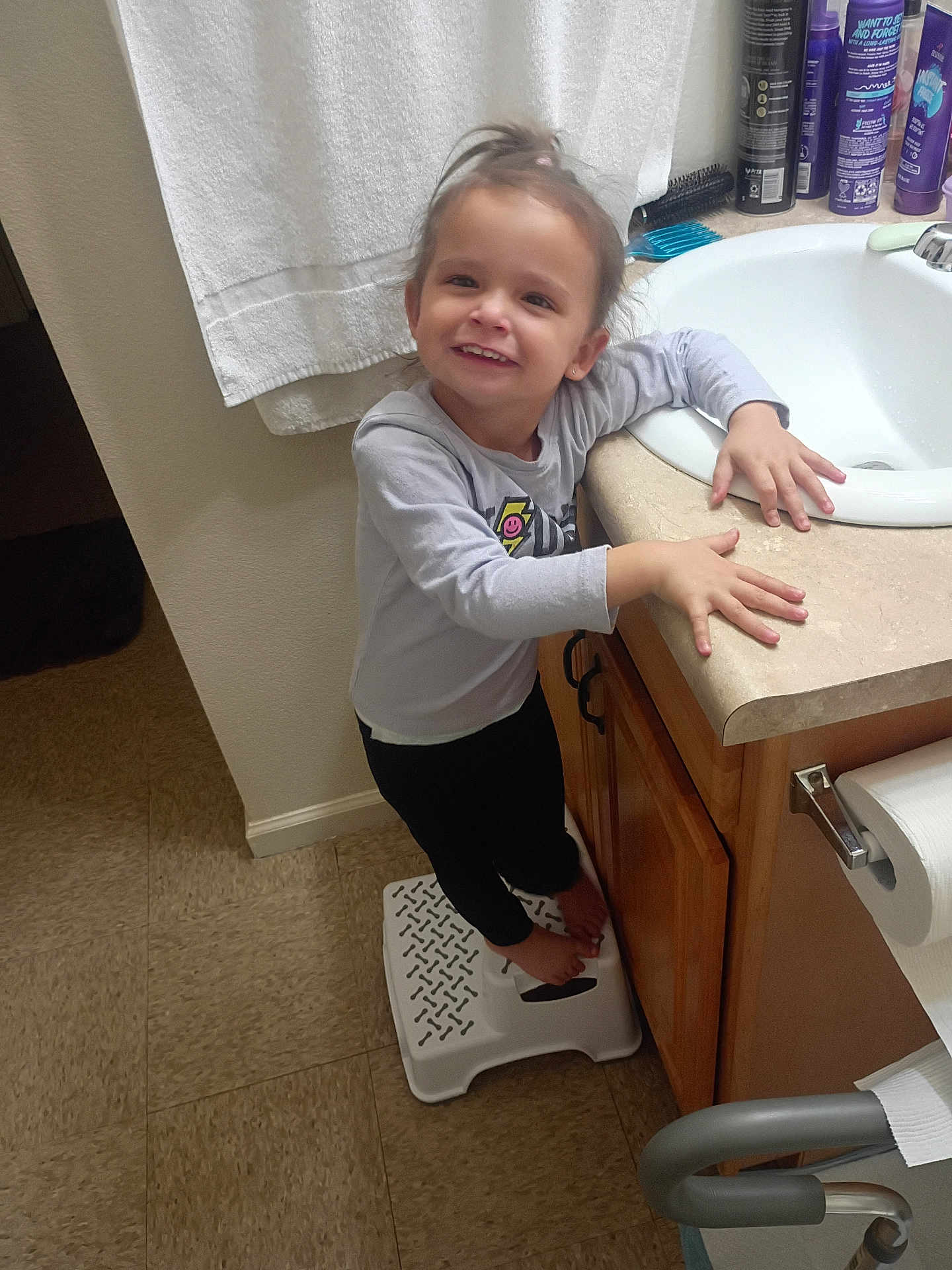 Madison is registered to the contest to win money with this photo: child, smiling, bathroom, sink, step_stool, towel, toiletries, countertop, floor, cabinet, person, barefoot, long_sleeve_shirt, black_pants, hair_tie, happy, indoor, young_child, standing, holding