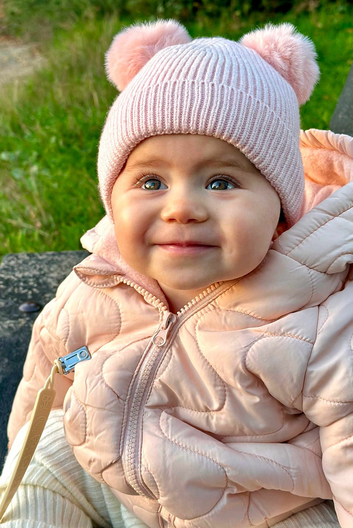 Amelia joined the competition — help win amazing prizes! baby, child, pink_hat, knit_hat, pom_poms, quilted_jacket, smile, outdoor, green_background, cute, warm_clothing, faces, portrait, happy, winter_wear, infant, closeup, baby_jacket, nature, eyes