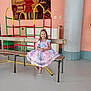 child, girl, dress, bench, indoor, playground, climbing_structure, smile, happy, floor, wall, artwork, colorful, footwear, sandals, hair_bow, radiator, metal, wood, person