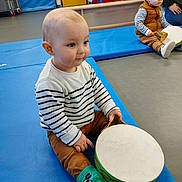 Lenny participe au concours pour gagner de l'argent avec cette photo : toddler, child, drum, striped_sweater, green_socks, indoor, playroom, gym_mat, floor, toy, musical_instrument, brown_pants, stacked_mats, yellow_door, curious, seated, play, baby, striped_clothing, person